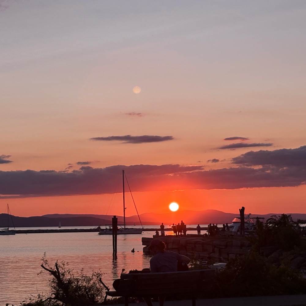 People on the waterfront docks watching the sun set over Lake Champlain, a sailboat in the harbor